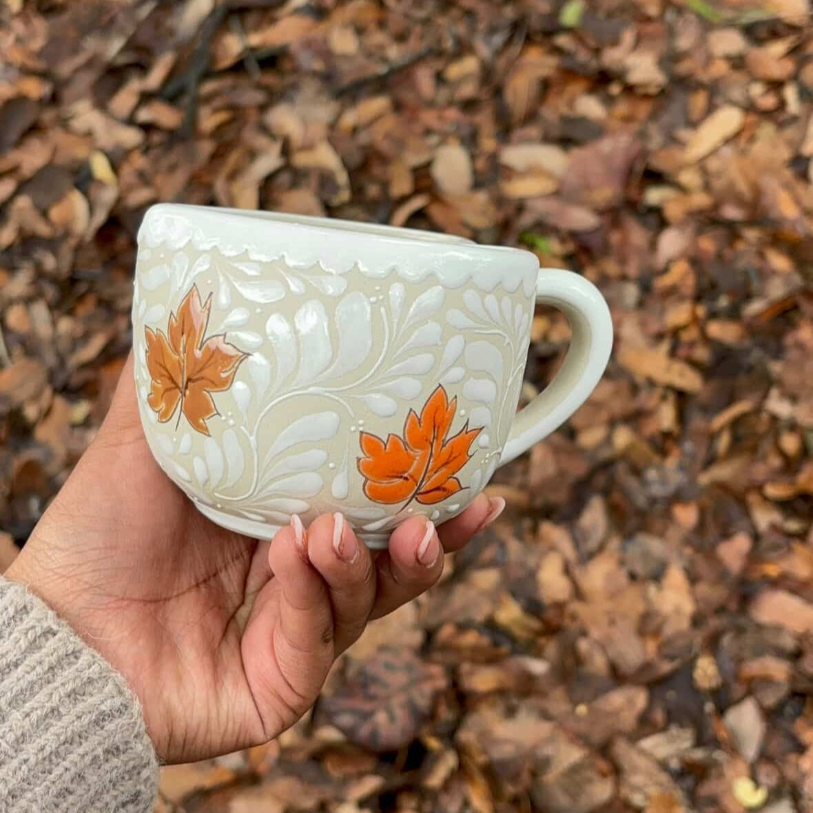 Hand holding a decorative teacup with autumn leaf design against a background of fallen leaves.